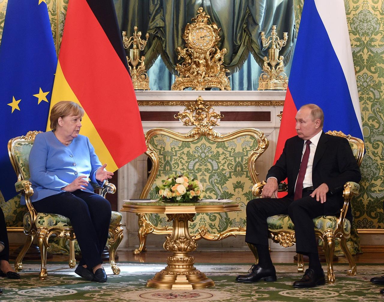 Angela Merkel und Wladimir Putin sitzen einander gegenüber auf Sesseln mit goldenen Ornamenten, hinter ihnen die Flaggen der EU, Deutschlands und Russlands.