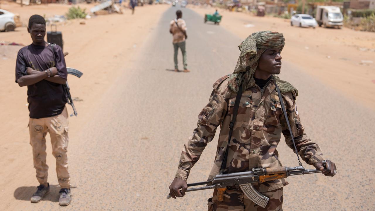Bewaffnete Personen sind auf einer Straße in Sudan zu sehen. Hinter ihnen Fahrzeuge und Gebäude.