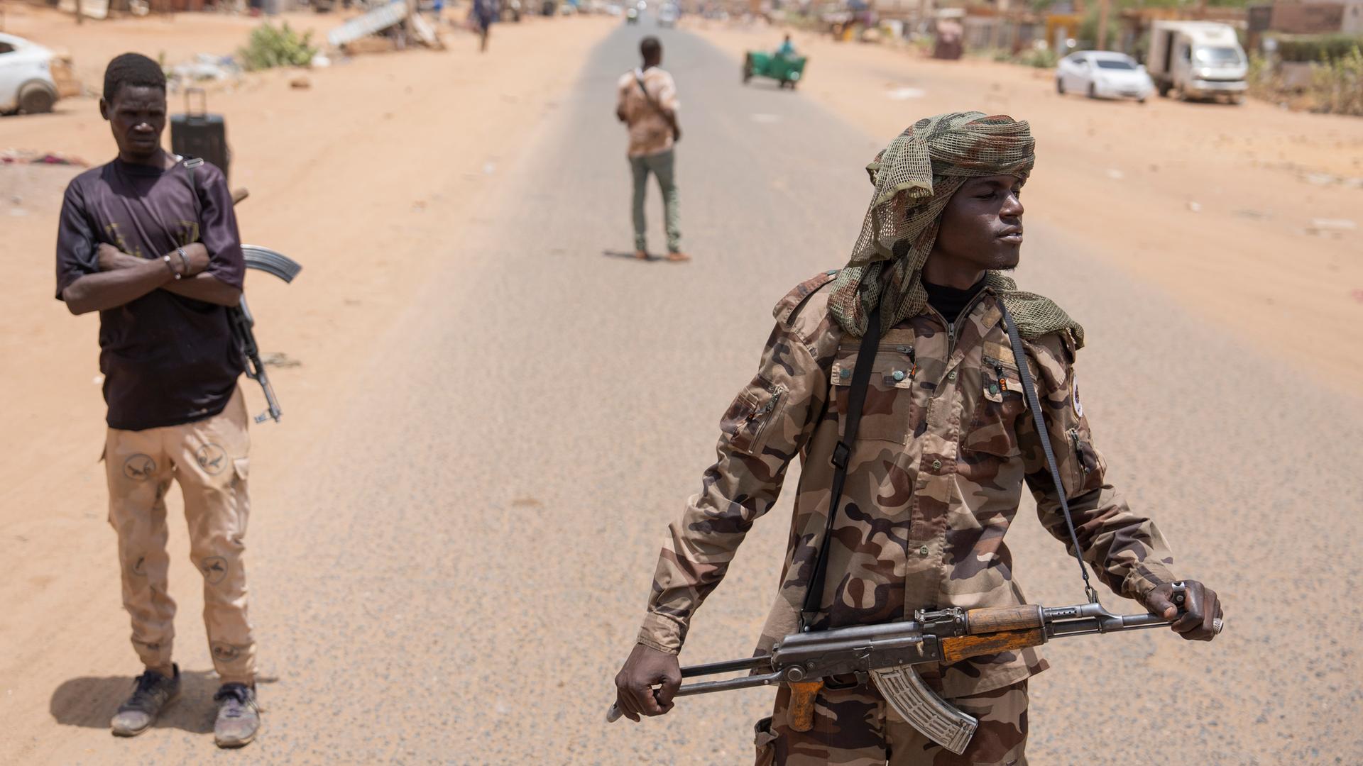 Bewaffnete Personen sind auf einer Straße in Sudan zu sehen. Hinter ihnen Fahrzeuge und Gebäude. Bewaffnete Personen sind auf einer Straße in Sudan zu sehen. Hinter ihnen Fahrzeuge und Gebäude.