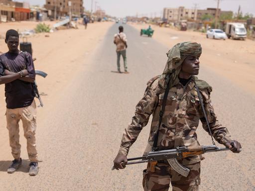 Bewaffnete Personen sind auf einer Straße in Sudan zu sehen. Hinter ihnen Fahrzeuge und Gebäude. Bewaffnete Personen sind auf einer Straße in Sudan zu sehen. Hinter ihnen Fahrzeuge und Gebäude.