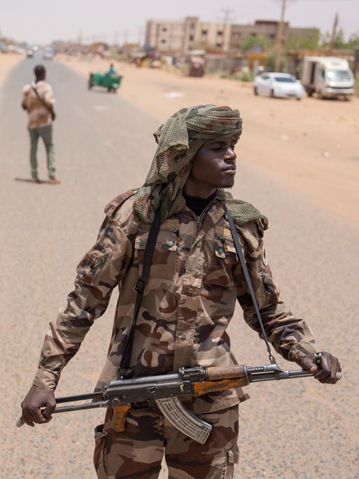 Bewaffnete Personen sind auf einer Straße in Sudan zu sehen. Hinter ihnen Fahrzeuge und Gebäude. Bewaffnete Personen sind auf einer Straße in Sudan zu sehen. Hinter ihnen Fahrzeuge und Gebäude.