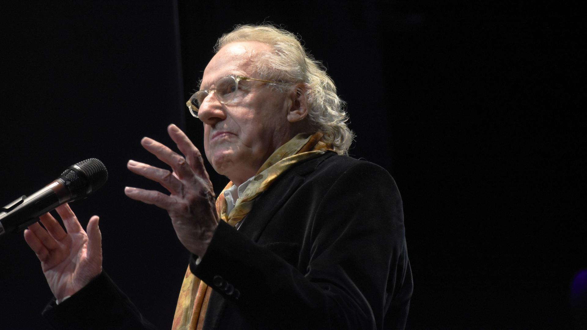 Walter Hess spricht auf einer Bühne in ein Mikrofon. Er trägt einen bunten Schal zum dunklen Sakko und eine transparent gefasste Brille.