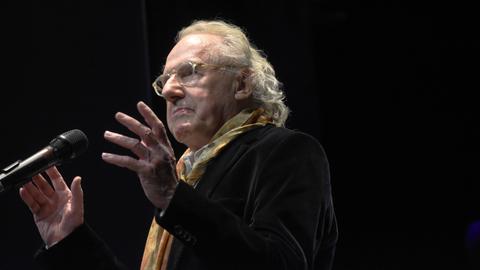 Walter Hess spricht auf einer Bühne in ein Mikrofon. Er trägt einen bunten Schal zum dunklen Sakko und eine transparent gefasste Brille. Walter Hess spricht auf einer Bühne in ein Mikrofon. Er trägt einen bunten Schal zum dunklen Sakko und eine transparent gefasste Brille.