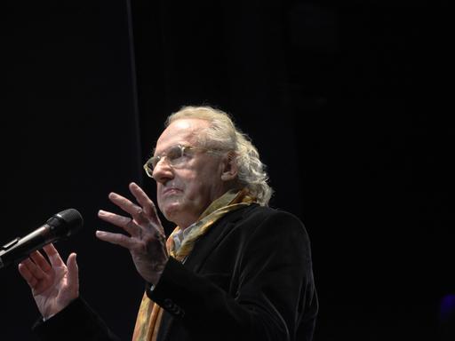 Walter Hess spricht auf einer Bühne in ein Mikrofon. Er trägt einen bunten Schal zum dunklen Sakko und eine transparent gefasste Brille.