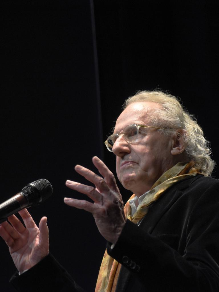 Walter Hess spricht auf einer Bühne in ein Mikrofon. Er trägt einen bunten Schal zum dunklen Sakko und eine transparent gefasste Brille.