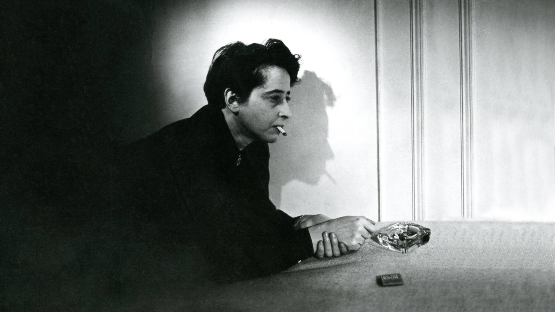 Filmstill aus "Hannah Arendt - Die Pflicht zum Ungehorsam" in dem Hannah Arendt Zigarette rauchend auf einem Tisch lehnt. Der Schatten ihres Profil ist an der Wand zu sehen.
