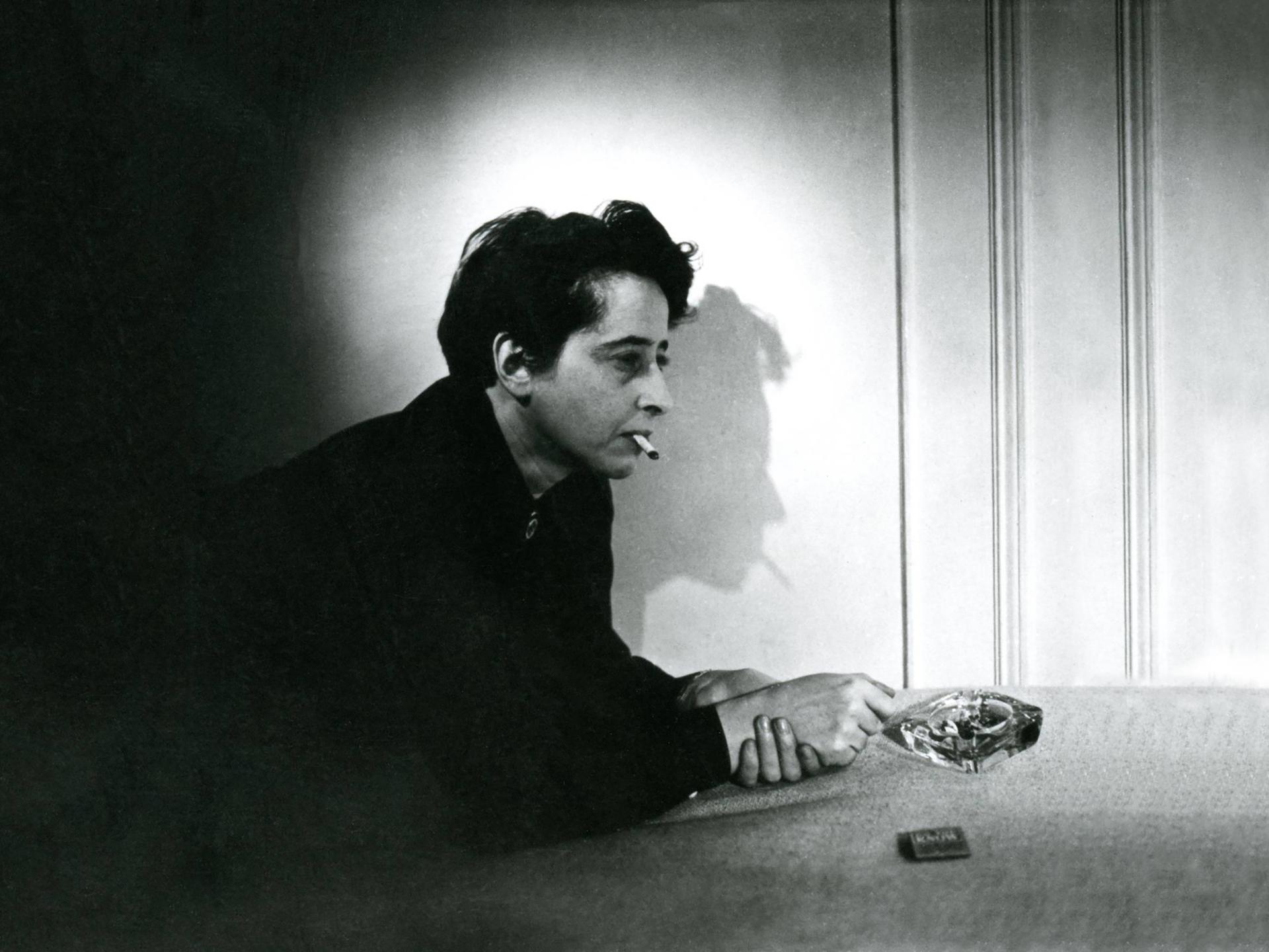 Filmstill aus "Hannah Arendt - Die Pflicht zum Ungehorsam" in dem Hannah Arendt Zigarette rauchend auf einem Tisch lehnt. Der Schatten ihres Profil ist an der Wand zu sehen.