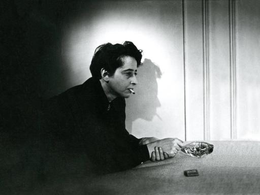 Filmstill aus "Hannah Arendt - Die Pflicht zum Ungehorsam" in dem Hannah Arendt Zigarette rauchend auf einem Tisch lehnt. Der Schatten ihres Profil ist an der Wand zu sehen.
