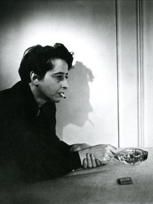 Filmstill aus "Hannah Arendt - Die Pflicht zum Ungehorsam" in dem Hannah Arendt Zigarette rauchend auf einem Tisch lehnt. Der Schatten ihres Profil ist an der Wand zu sehen.