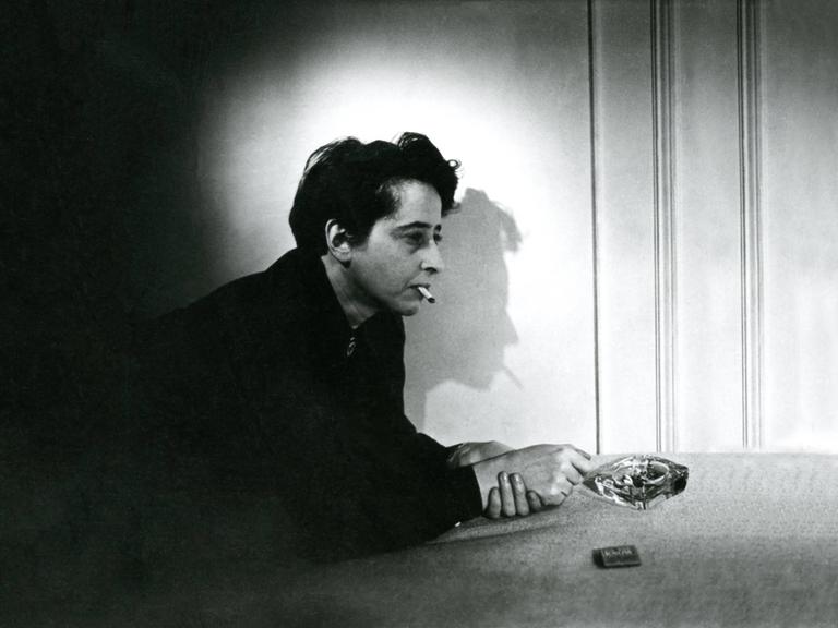 Filmstill aus "Hannah Arendt - Die Pflicht zum Ungehorsam" in dem Hannah Arendt Zigarette rauchend auf einem Tisch lehnt. Der Schatten ihres Profil ist an der Wand zu sehen.