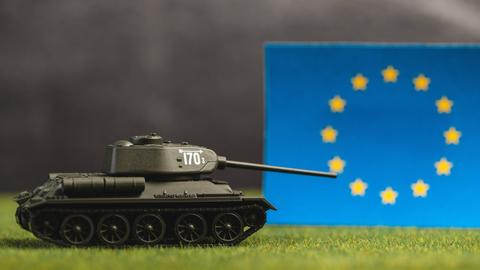 Ein Spielzeugpanzer mit gesenktem Schussrohr vor einer EU-Flagge und einem düsteren undeutlichen Hintergrund Ein Spielzeugpanzer mit gesenktem Schussrohr vor einer EU-Flagge und einem düsteren undeutlichen Hintergrund