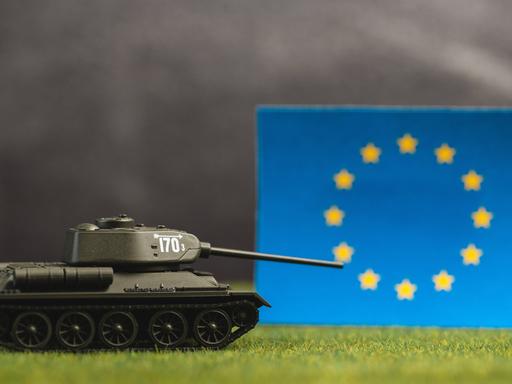 Ein Spielzeugpanzer mit gesenktem Schussrohr vor einer EU-Flagge und einem düsteren undeutlichen Hintergrund
