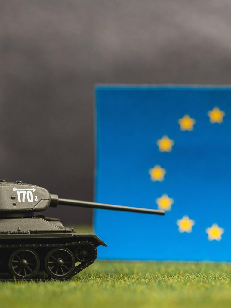 Ein Spielzeugpanzer mit gesenktem Schussrohr vor einer EU-Flagge und einem düsteren undeutlichen Hintergrund