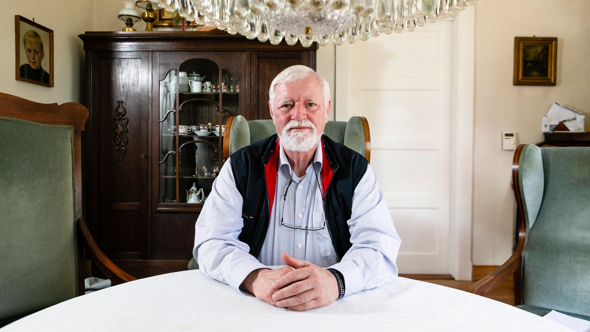 Bürgermeister-Macher Thies Thiessen sitzt auf einem Ohrensessel in seinem Wohnhaus. Er trägt weiße Haare und einen weißen Bart. Seine Hände liegen auf dem weißen Tisch. Über ihm sieht man einen Kronleuchter. Im Hintergrund erkennt man einen alten Schrank und zwe Gemälde.