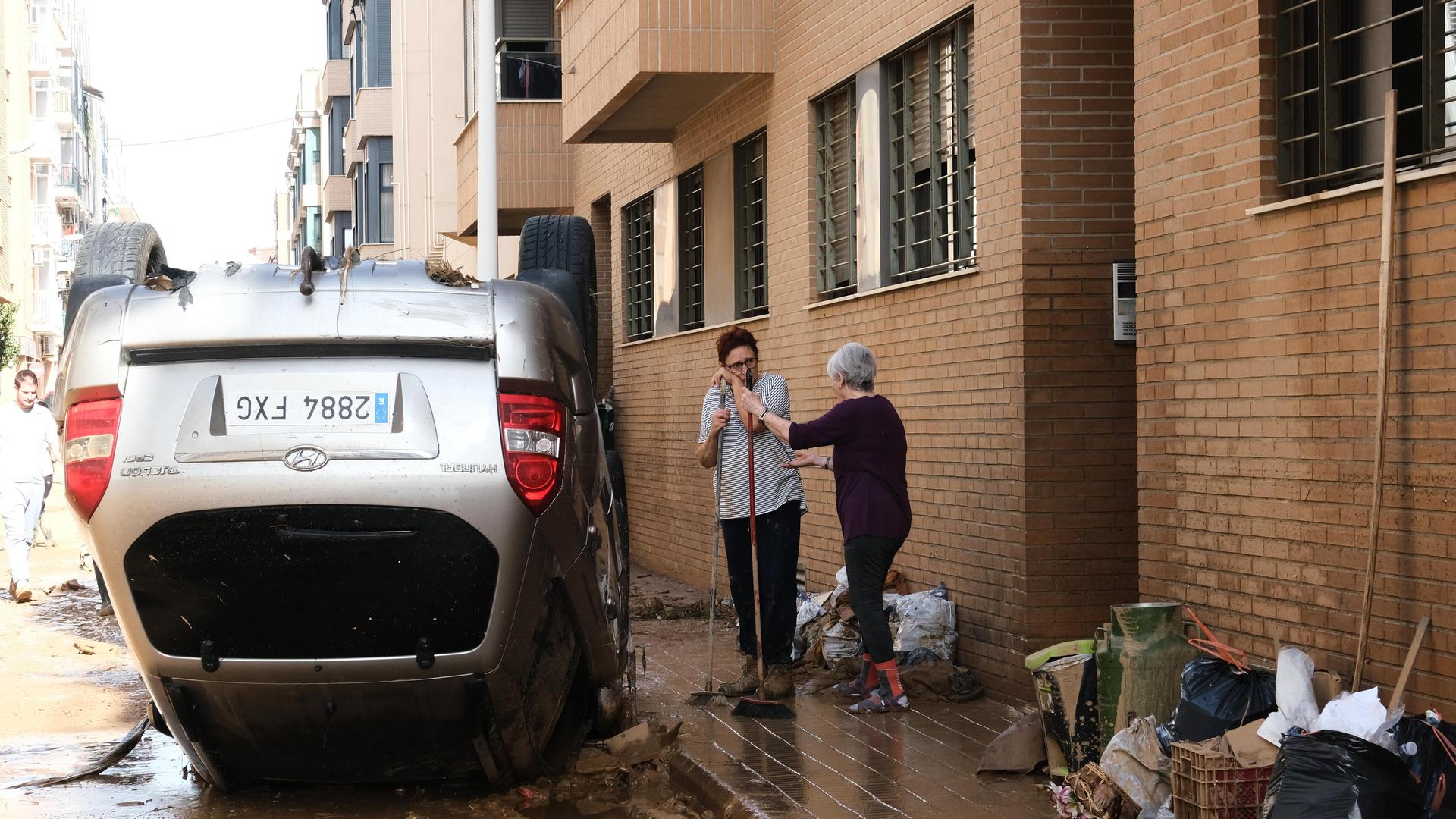 Zwei Frauen fegen vor einer Haustür nach einem schweren Unwetter in Spanien, Neben ihnen liegt ein Auto auf dem Dach.