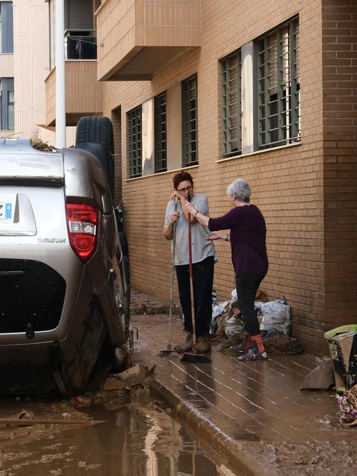 Zwei Frauen fegen vor einer Haustür nach einem schweren Unwetter in Spanien, Neben ihnen liegt ein Auto auf dem Dach.