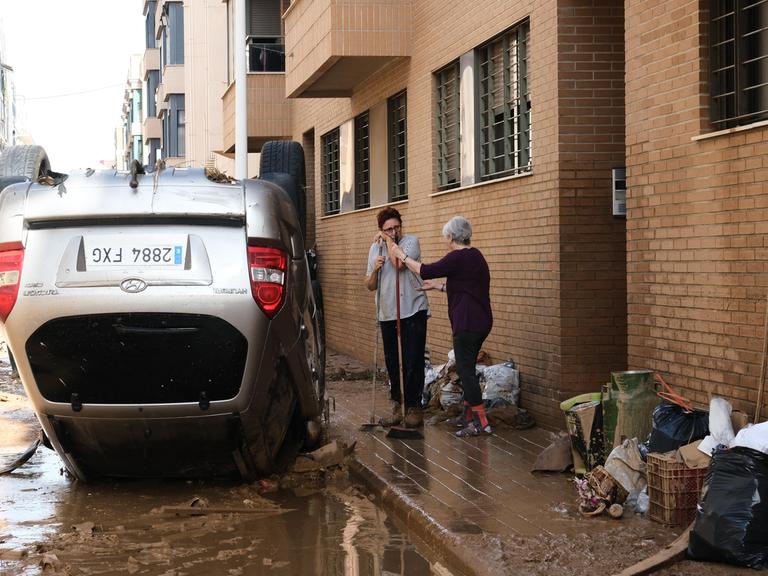 Zwei Frauen fegen vor einer Haustür nach einem schweren Unwetter in Spanien, Neben ihnen liegt ein Auto auf dem Dach.