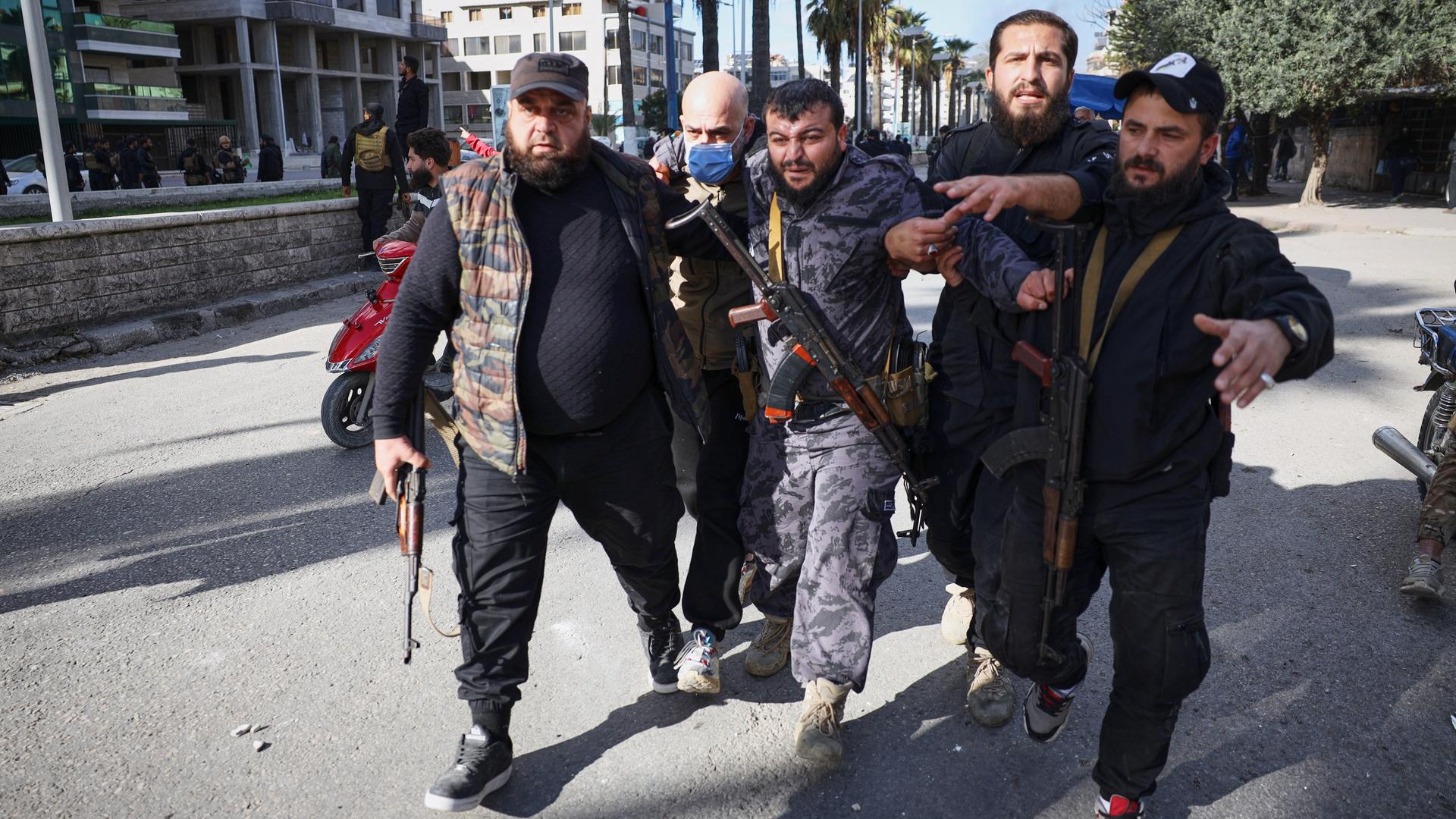 Auf einer Straße mit hohen Gebäuden stützen mehrere bewaffnete Männer den Verletzten in ihrer Mitte. Auf einer Straße mit hohen Gebäuden stützen mehrere bewaffnete Männer den Verletzten in ihrer Mitte.