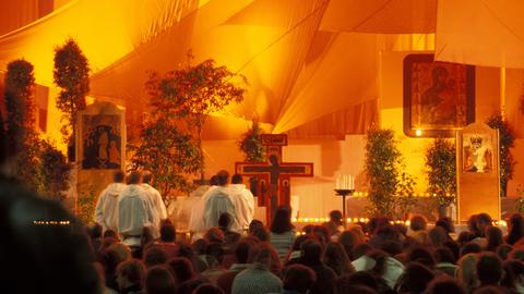 Ein orange beleuchteter Raum ist gefüllt mit Menschen. Im Vordergrund sind Männer mit weißen Roben zu sehen und Jesus am Kreuz