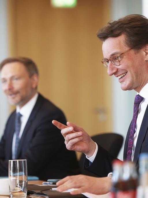 Zwei Männer in Anzügen sitzen an einem Tisch mit Kaffeekannen und Gläsern.