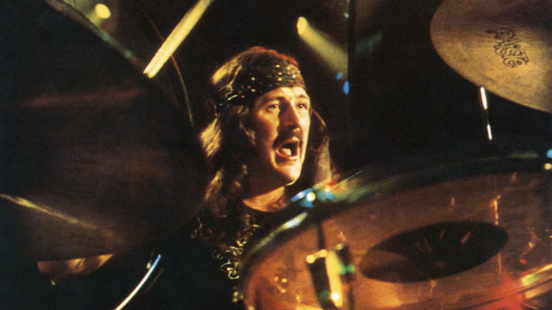 John Bonham spielt Schlagzeug, Im Vordergrund sind einige Bestandteile des Schlagzeugs zu sehen, dahinter Bonham im Alter von rund 30 Jahren. Er trägt die Haare lang, ein Stirnband und hat einen Oberlippenbart.