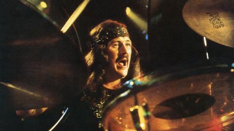 John Bonham spielt Schlagzeug, Im Vordergrund sind einige Bestandteile des Schlagzeugs zu sehen, dahinter Bonham im Alter von rund 30 Jahren. Er trägt die Haare lang, ein Stirnband und hat einen Oberlippenbart.