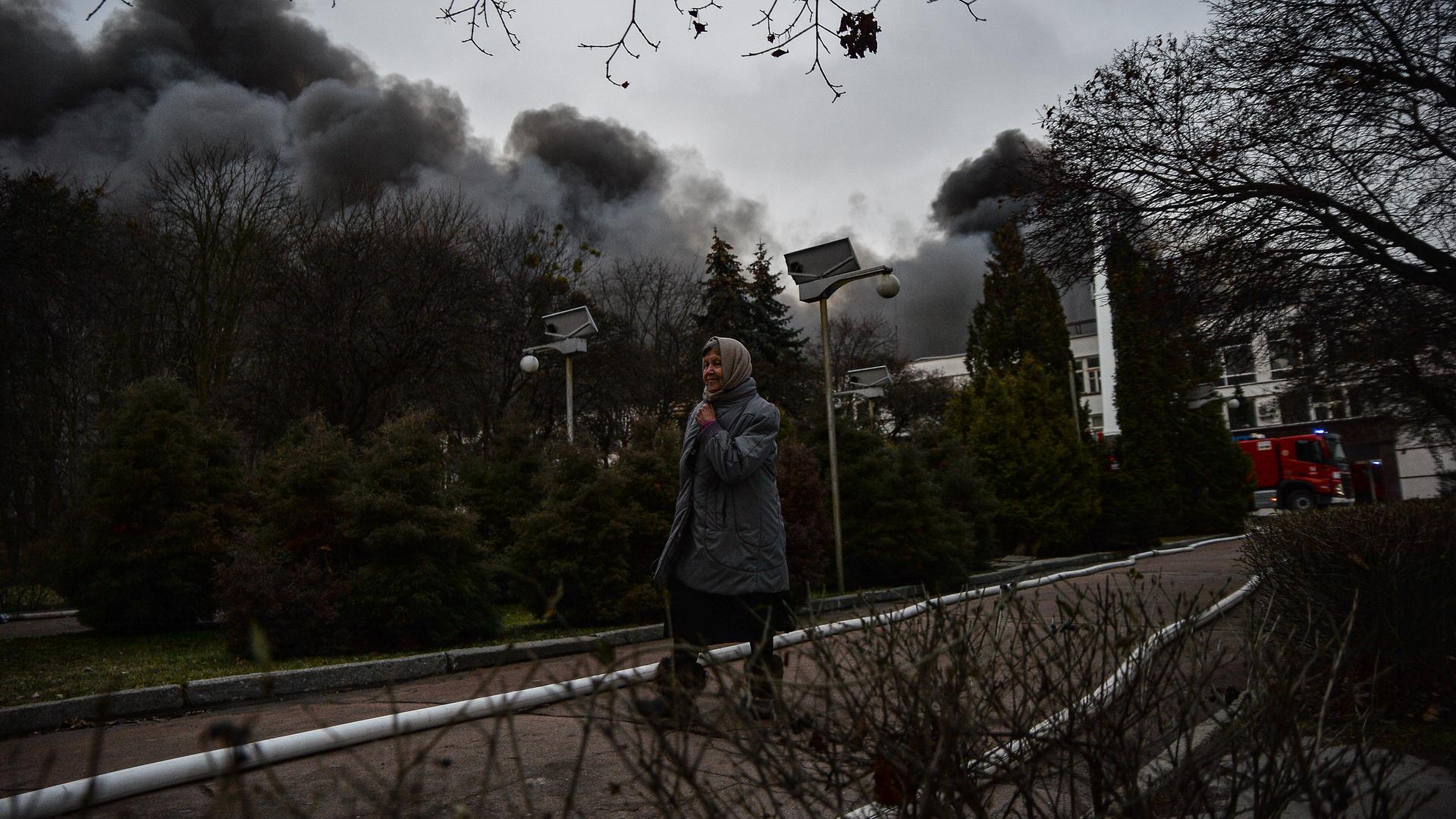 Eine ältere Frau läuft auf einer Straße in Kiew, im Hintergrund steigt die Rauchsäule einer zerstörten Fabrik auf.
