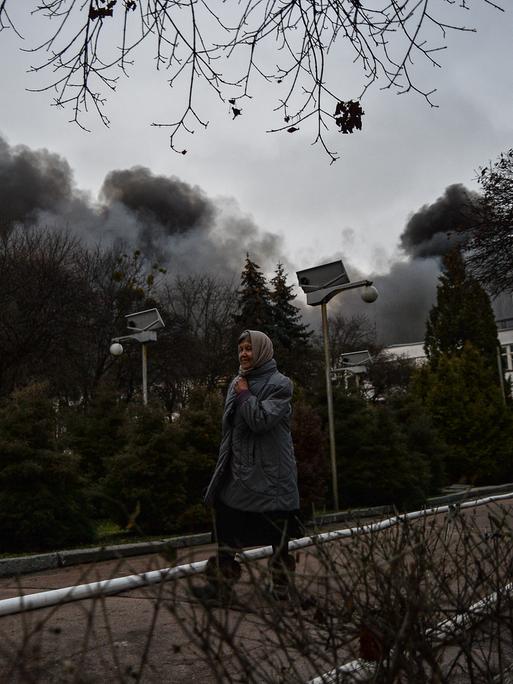 Eine ältere Frau läuft auf einer Straße in Kiew, im Hintergrund steigt die Rauchsäule einer zerstörten Fabrik auf.