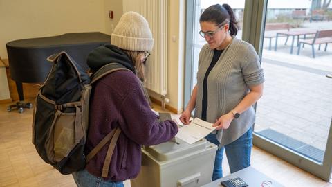 Eine junge Frau wirft ihren Wahlzettel in eine Wahlurne. Eine andere Frau beobachtet das.  