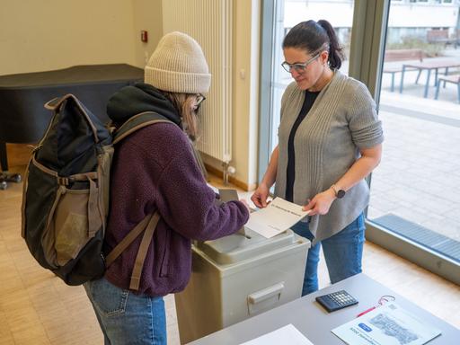Eine junge Frau wirft ihren Wahlzettel in eine Wahlurne. Eine andere Frau beobachtet das.  