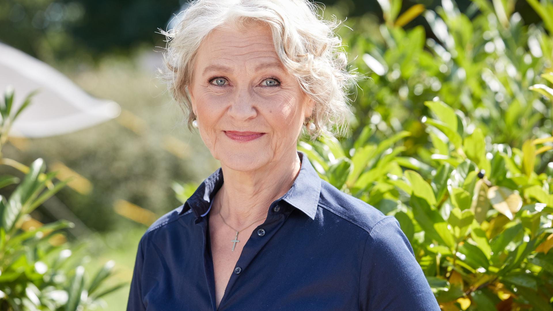Edelgard Hansen trägt eine blaue Bluse, steht vor einem Busch und schaut freundlich in die Kamera.