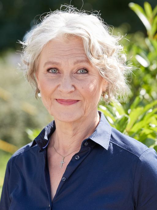 Edelgard Hansen trägt eine blaue Bluse, steht vor einem Busch und schaut freundlich in die Kamera.
