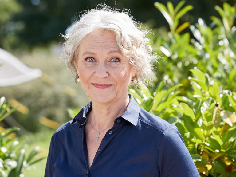 Edelgard Hansen trägt eine blaue Bluse, steht vor einem Busch und schaut freundlich in die Kamera.