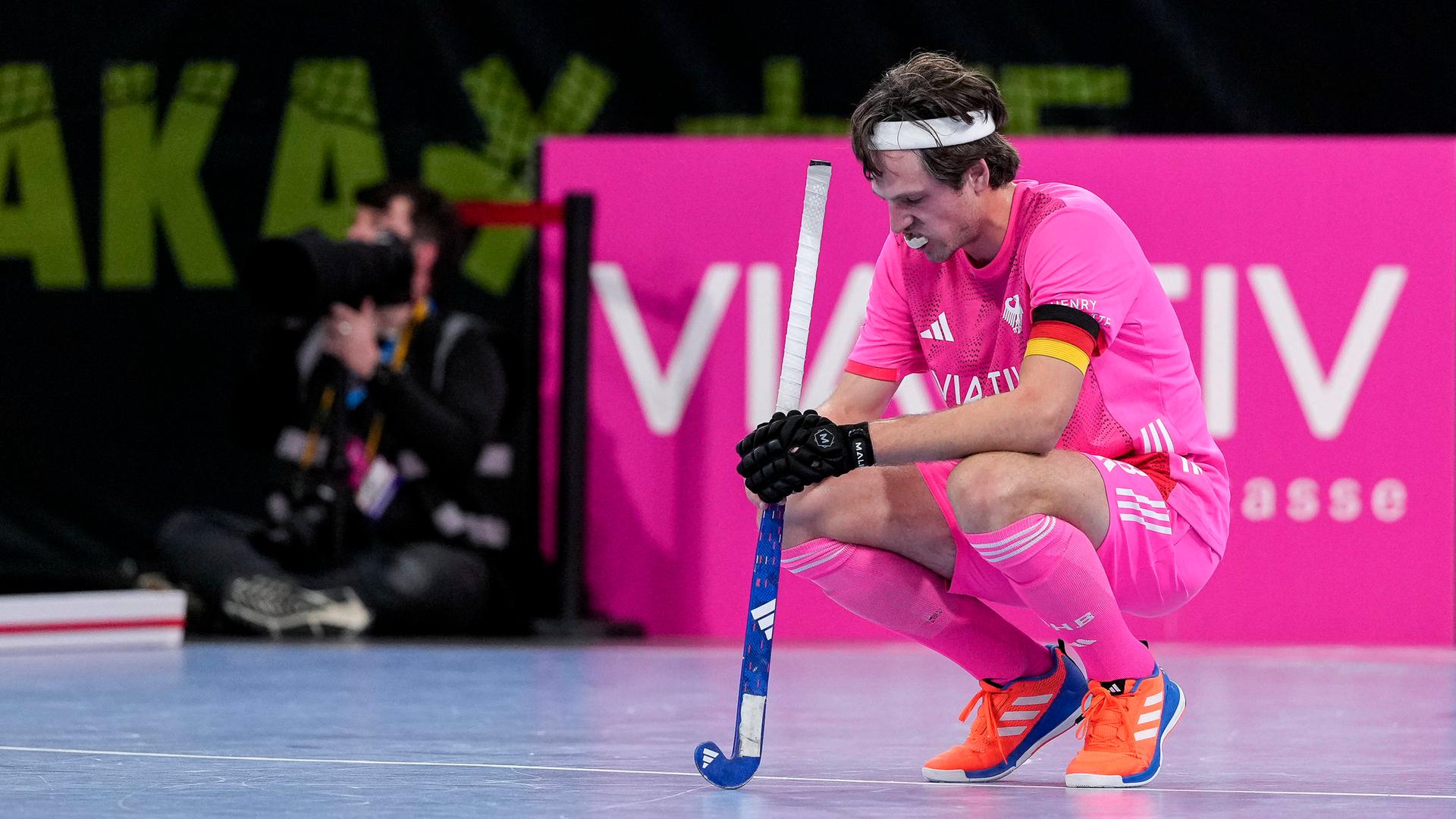 Ein Hockeyspieler in pinkem Trikot und mit Kapitänsbinde in Deutschland-Farben hocket enttäucht auf dem Boden einer Sporthalle. Er hat einen Hockey-Schläger in der Hand.
