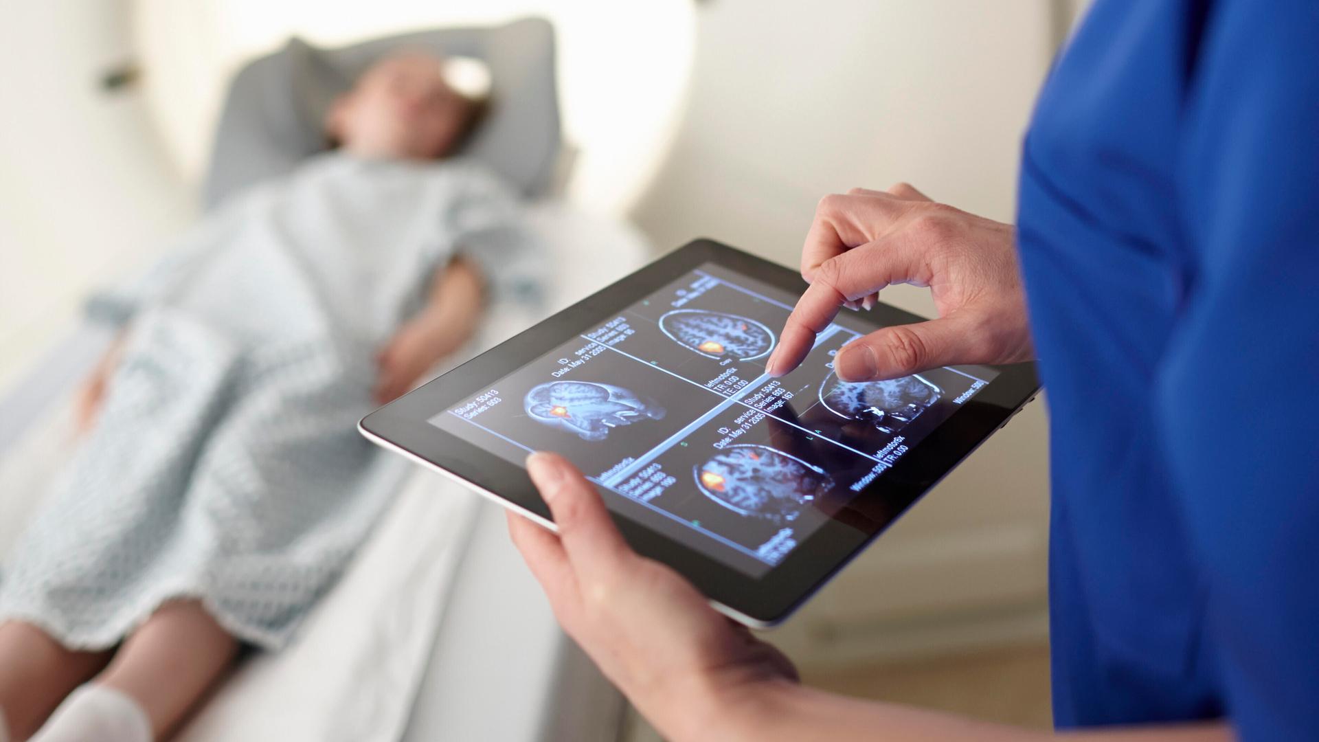Eine Frau betrachtet Scans des Gehirns auf einem Tablet. Im Hintergrund liegt ein Kind bei der Untersuchung in einem radiologischen Apparat auf einer Liege. Eine Frau betrachtet Scans des Gehirns auf einem Tablet. Im Hintergrund liegt ein Kind bei der Untersuchung in einem radiologischen Apparat auf einer Liege.