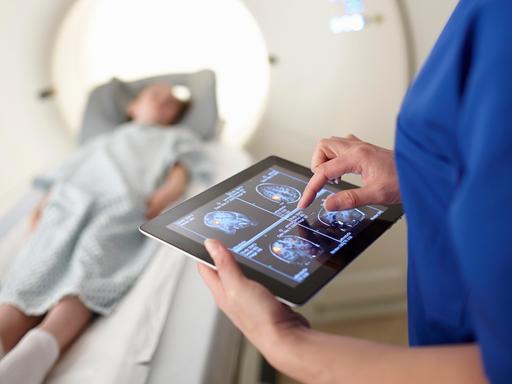 Eine Frau betrachtet Scans des Gehirns auf einem Tablet. Im Hintergrund liegt ein Kind bei der Untersuchung in einem radiologischen Apparat auf einer Liege.