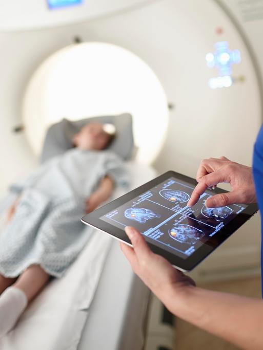 Eine Frau betrachtet Scans des Gehirns auf einem Tablet. Im Hintergrund liegt ein Kind bei der Untersuchung in einem radiologischen Apparat auf einer Liege.