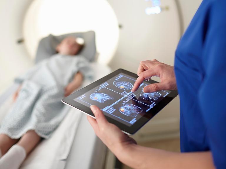 Eine Frau betrachtet Scans des Gehirns auf einem Tablet. Im Hintergrund liegt ein Kind bei der Untersuchung in einem radiologischen Apparat auf einer Liege.