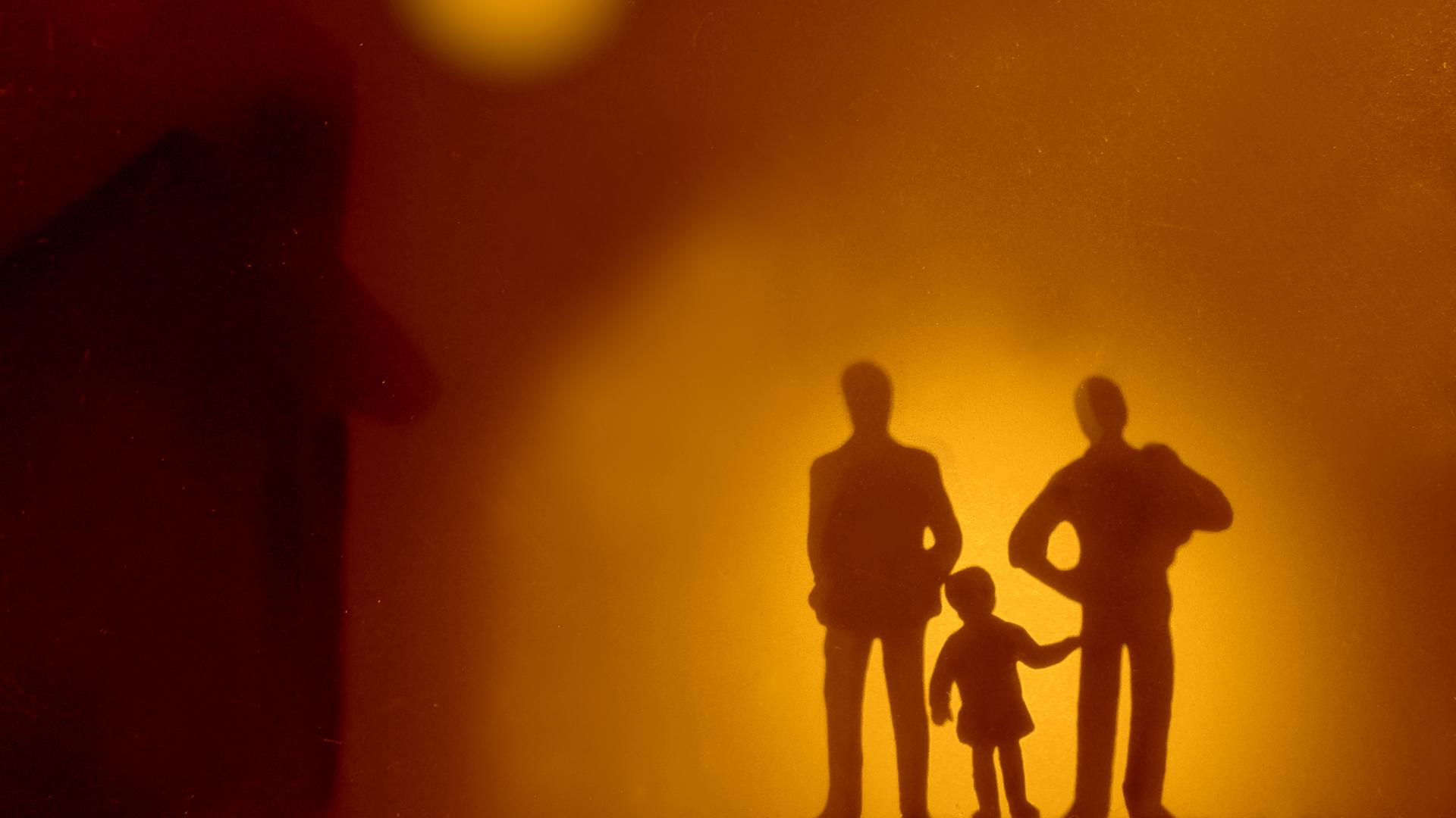 Die Silhouetten zweier Erwachsener, zwischen denen ein Kind steht. Die Silhouetten zweier Erwachsener, zwischen denen ein Kind steht.