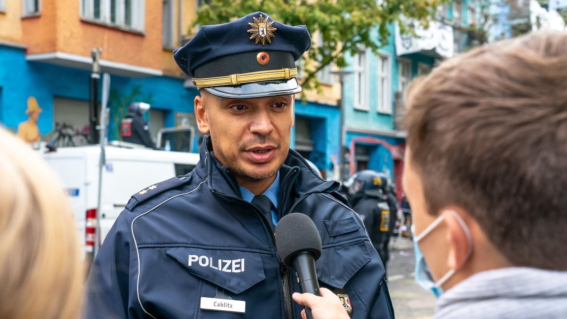 Thilo Calblitz in Polizeiuniform unf mit Polizeimütze auf dem Kopf spricht mit einem anderen Menschen, der ein Mikrofon in der Hand hält. Diese Person ist von hinten zu sehen.