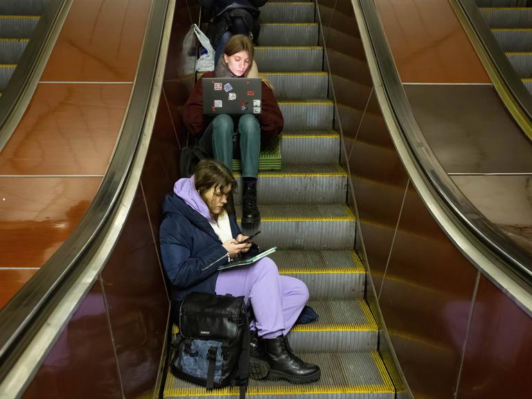 Junge Menschen mit Smartphone und Laptop sitzen auf einer Rolltreppe in einer U-Bahn Station in Kiew und warten einen Luftangriffalarm ab. 13. Dezember 2022, Kiew, Ukraine.