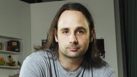 Porträt von Jan Costin Wagner. Er hat schulterlanges dunkelbraunes Haar und trägt einen hellgrauen Pulli.