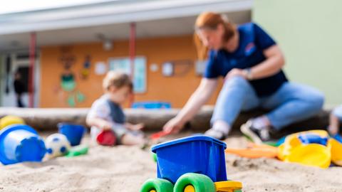 Ein Kind spielt mit einer Erzieherin im Sandkasten einer Kita. Im Vordergrund ist ein buntes Plastikspielzeug zu sehen.