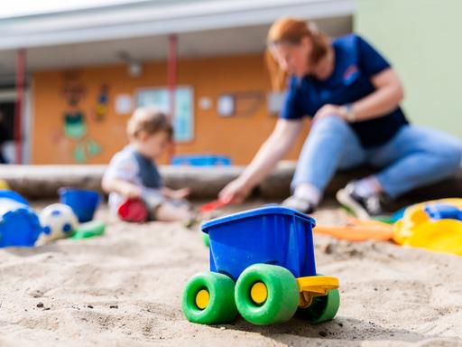 Ein Kind spielt mit einer Erzieherin im Sandkasten einer Kita. Im Vordergrund ist ein buntes Plastikspielzeug zu sehen.