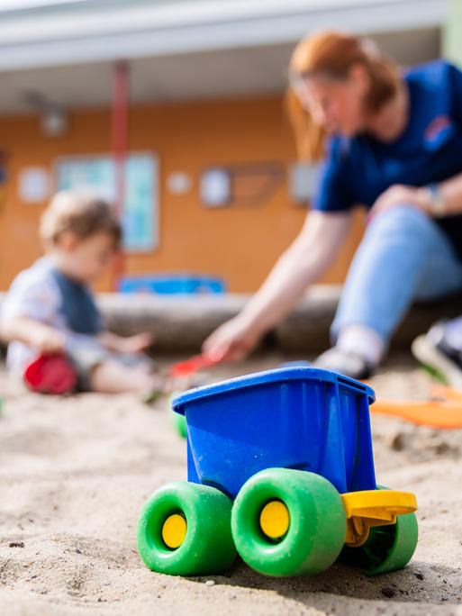 Ein Kind spielt mit einer Erzieherin im Sandkasten einer Kita. Im Vordergrund ist ein buntes Plastikspielzeug zu sehen.