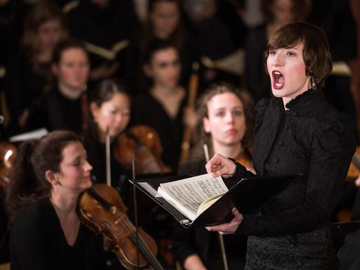 Eine Frau steht mit Noten in der Hand singend in einem Orchester