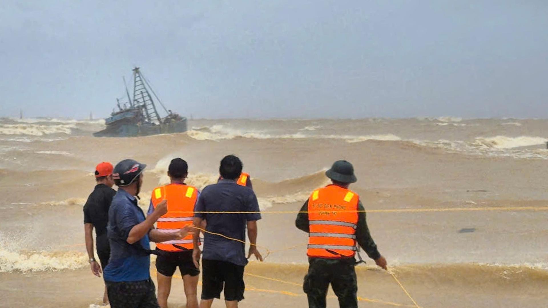 Man sieht die Rettungskräfte mit orangefarbenen Schwimmwesten von hinten. Im aufgepeitschten braunen Meer liegt schräg das Fischerboot.