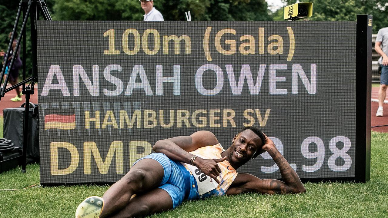 Der Leichtathlet Owen Ansah liegt auf einem Rasen vor einem Schild und zeigt einen Daumen hoch. Auf dem Schild ist zu lesen: 100m, Ansah Owen, Hamburger SV. 