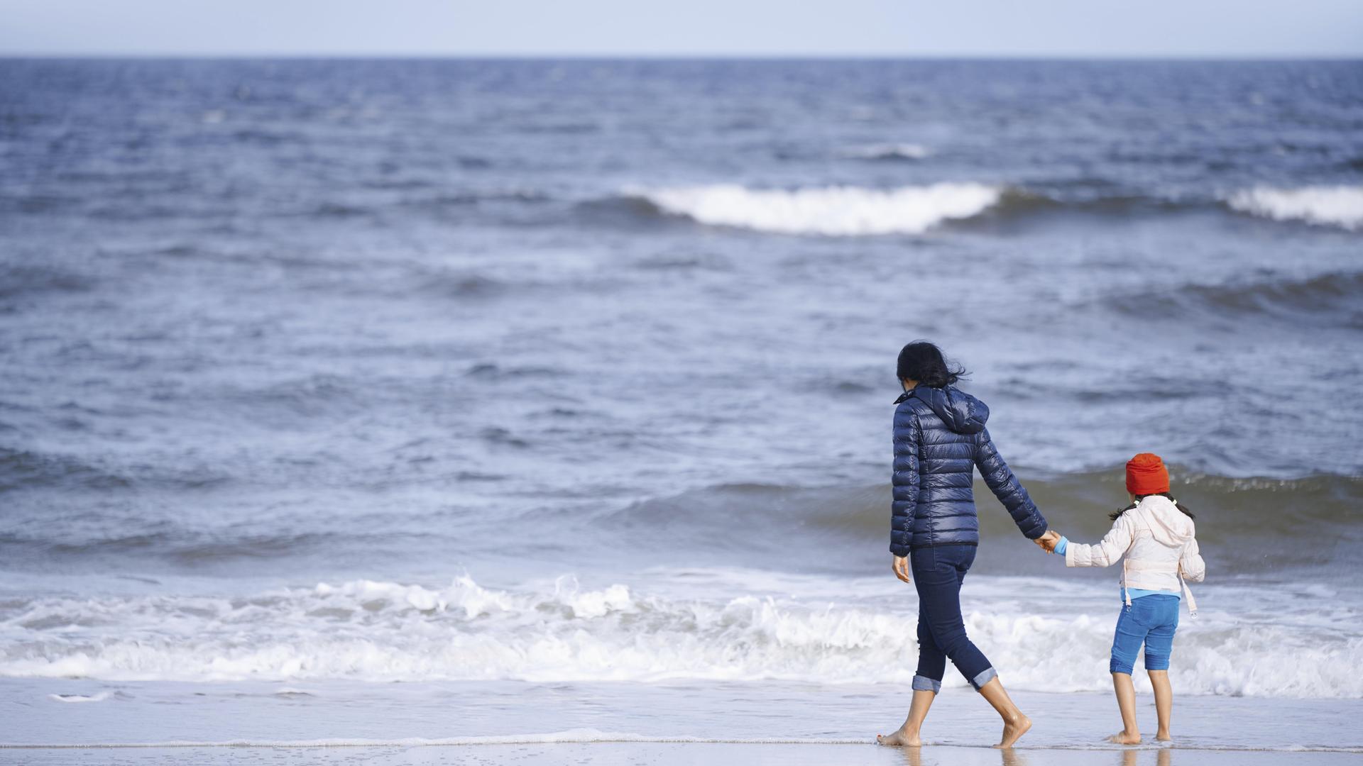 Ein Mutter hält ihre Tochter an der Hand und spaziert mit ihrem am Strand entlang. Im Hintergrund ist das weiter Meer zu sehen. Ein Mutter hält ihre Tochter an der Hand und spaziert mit ihrem am Strand entlang. Im Hintergrund ist das weiter Meer zu sehen.