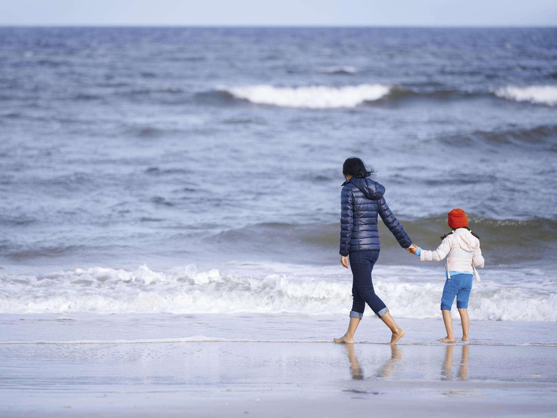 Ein Mutter hält ihre Tochter an der Hand und spaziert mit ihrem am Strand entlang. Im Hintergrund ist das weiter Meer zu sehen.